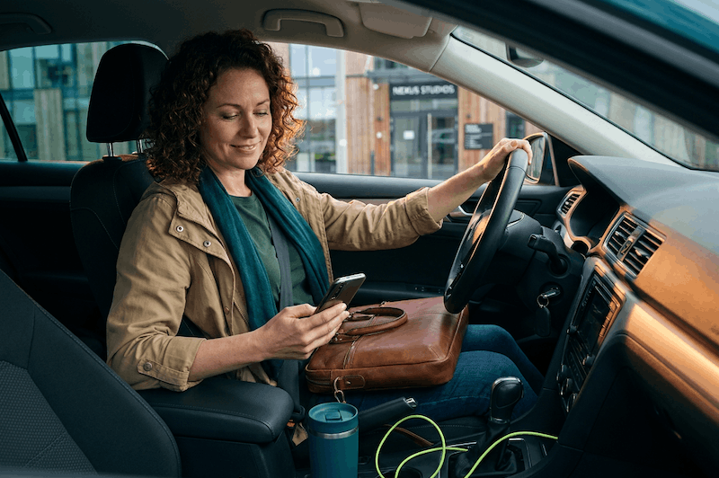 Freelancer woman in her early 40s seated in a parked car, looking at her phone with a confident soft smile while parked outside a client's modern office; warm natural daylight and shallow depth of field keep focus on her and the car interior.